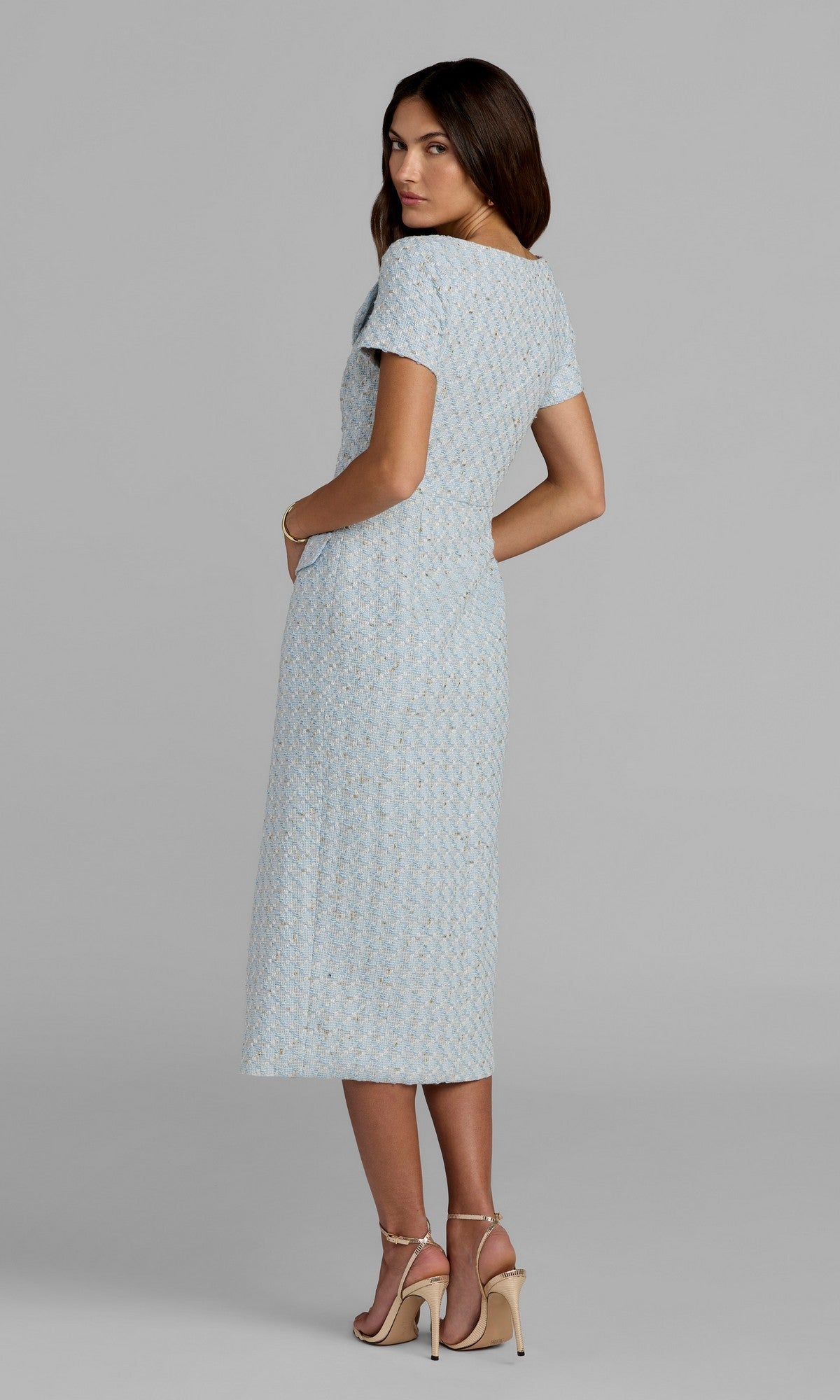 A woman with her back turned, looking over her shoulder, models the Mac Duggal 56374 Tea-Length Party Dress in light blue—a textured, short-sleeved style that falls to the knee—paired with beige high-heeled sandals against a plain background.